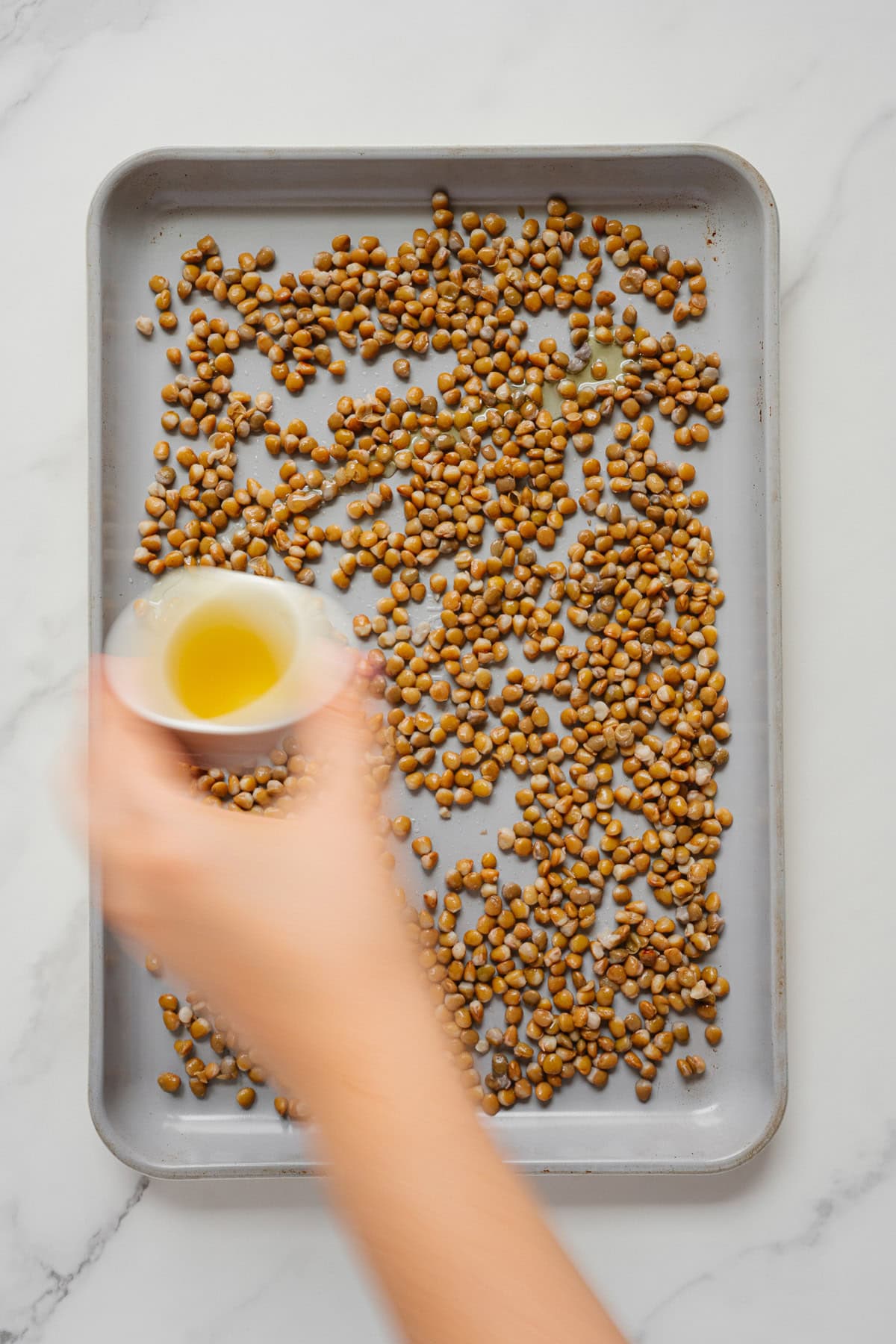 A hand pouring oil from a small glass into a baking tray filled with uncooked chickpeas and crispy lentils on a marble surface.