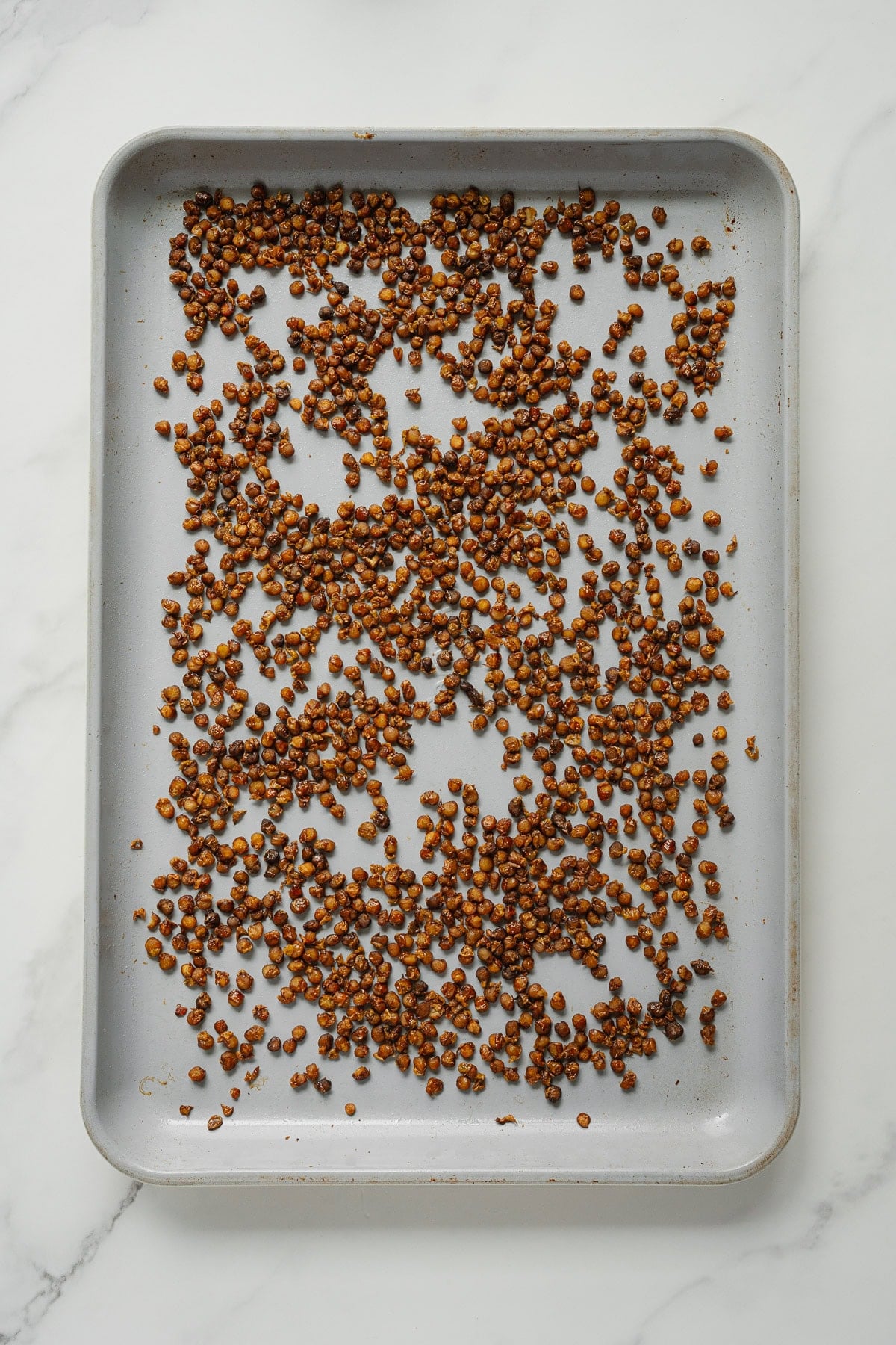 A baking sheet with a scattered layer of roasted, crispy chickpeas and crispy lentils on a white marble surface.