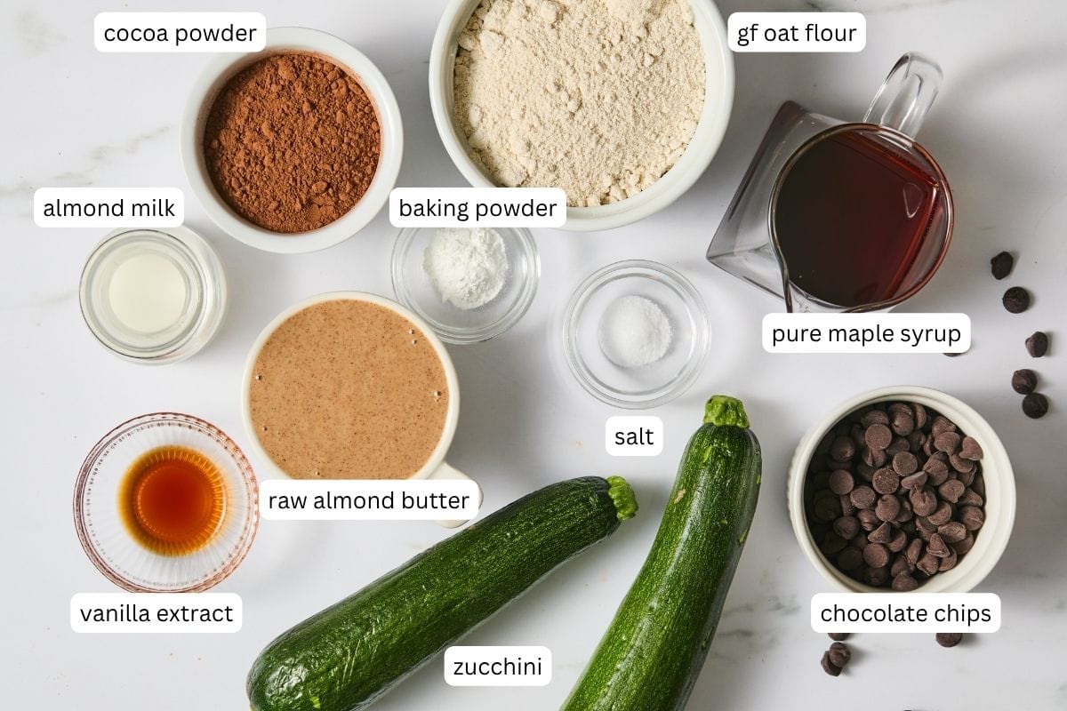 Ingredients for baking zucchini brownies are arranged on a white surface, each labeled: cocoa powder, almond milk, vanilla extract, raw almond butter, baking powder, oat flour, maple syrup, salt, zucchini, and chocolate chips.