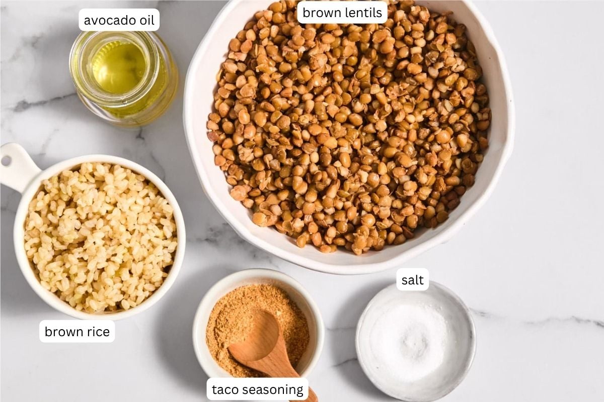 A bowl of cooked brown lentils for Lentil Taco Meat, a cup of brown rice, a small bowl of salt, a bowl of taco seasoning with a spoon, and a bottle of avocado oil on a white surface.