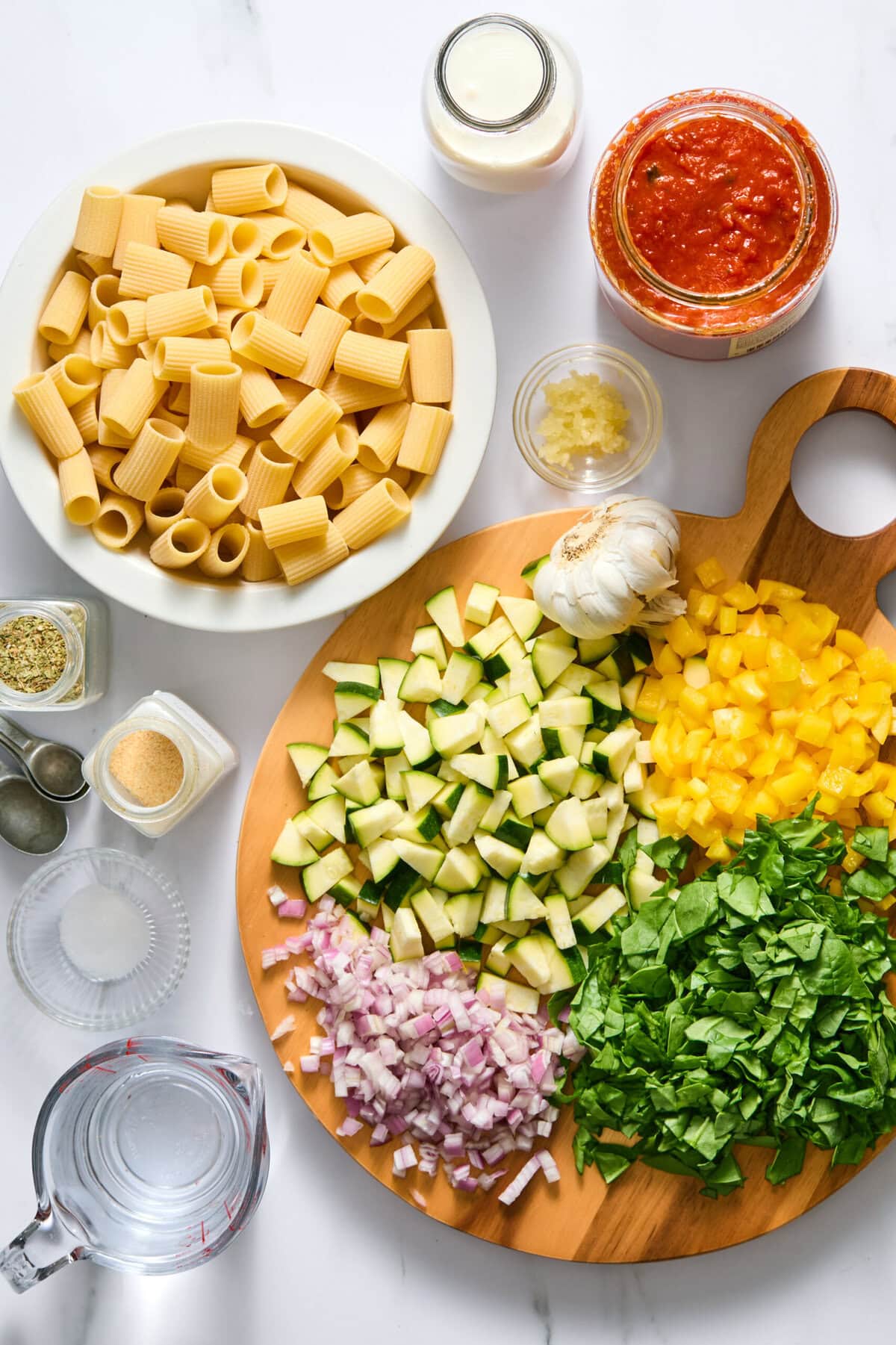 Sliced zucchini, yellow bell pepper, red onion, and spinach on a board, with uncooked pasta, tomato sauce, cream, spices, garlic, and a measuring cup of water arranged around them on a white surface.