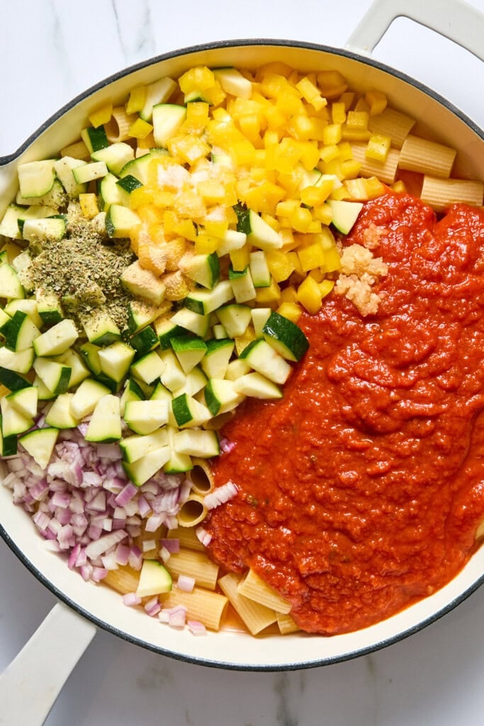 A large white pan with uncooked pasta, chopped zucchini, yellow bell pepper, red onion, dried herbs, minced garlic, and tomato sauce, all separated and ready to be mixed.