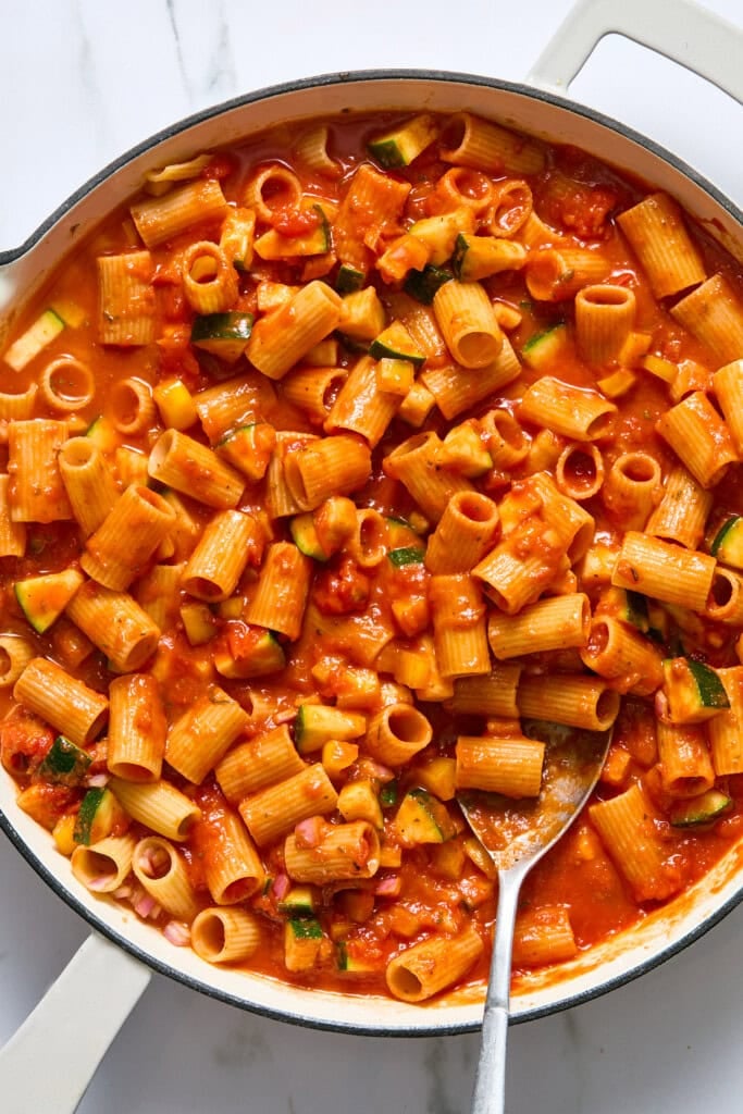 A pan filled with rigatoni pasta in a chunky tomato sauce with diced zucchini, stirred with a metal spoon.