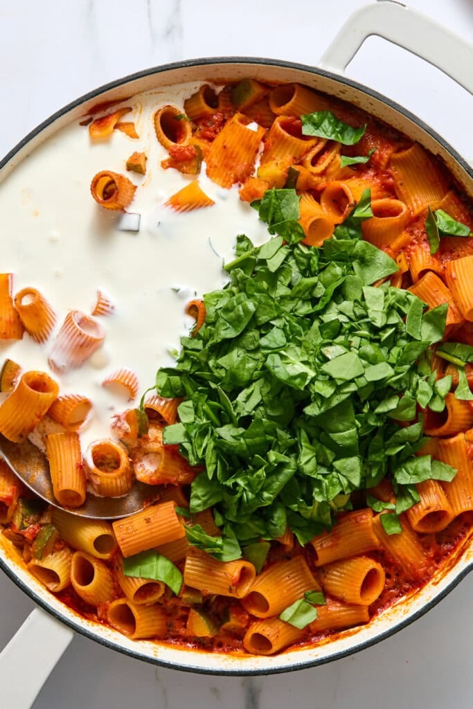 A skillet of rigatoni pasta in tomato sauce topped with chopped spinach and a splash of cream, ready to be mixed together.