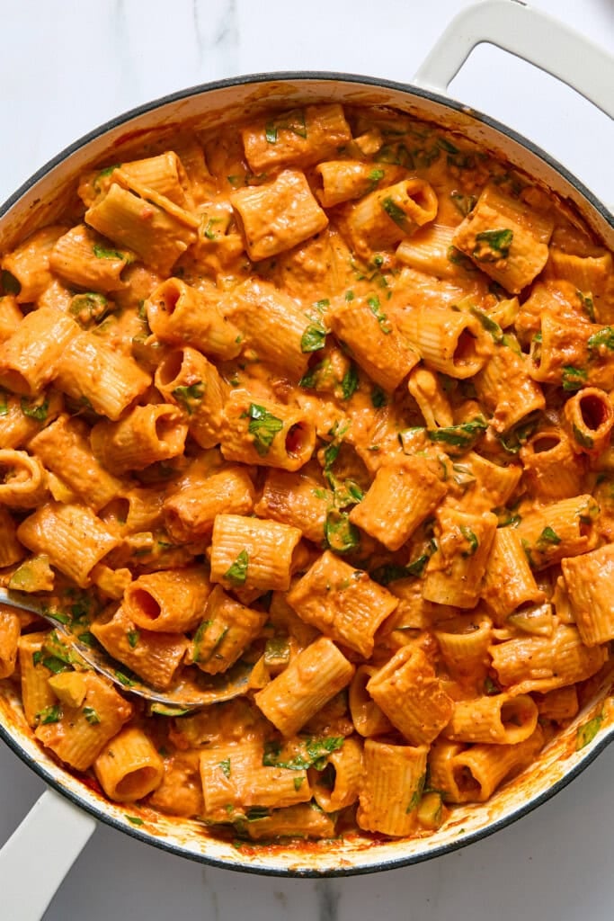 Large rigatoni pasta in a creamy, orange-tinted tomato sauce with green herbs, served in a white pan with a silver spoon.