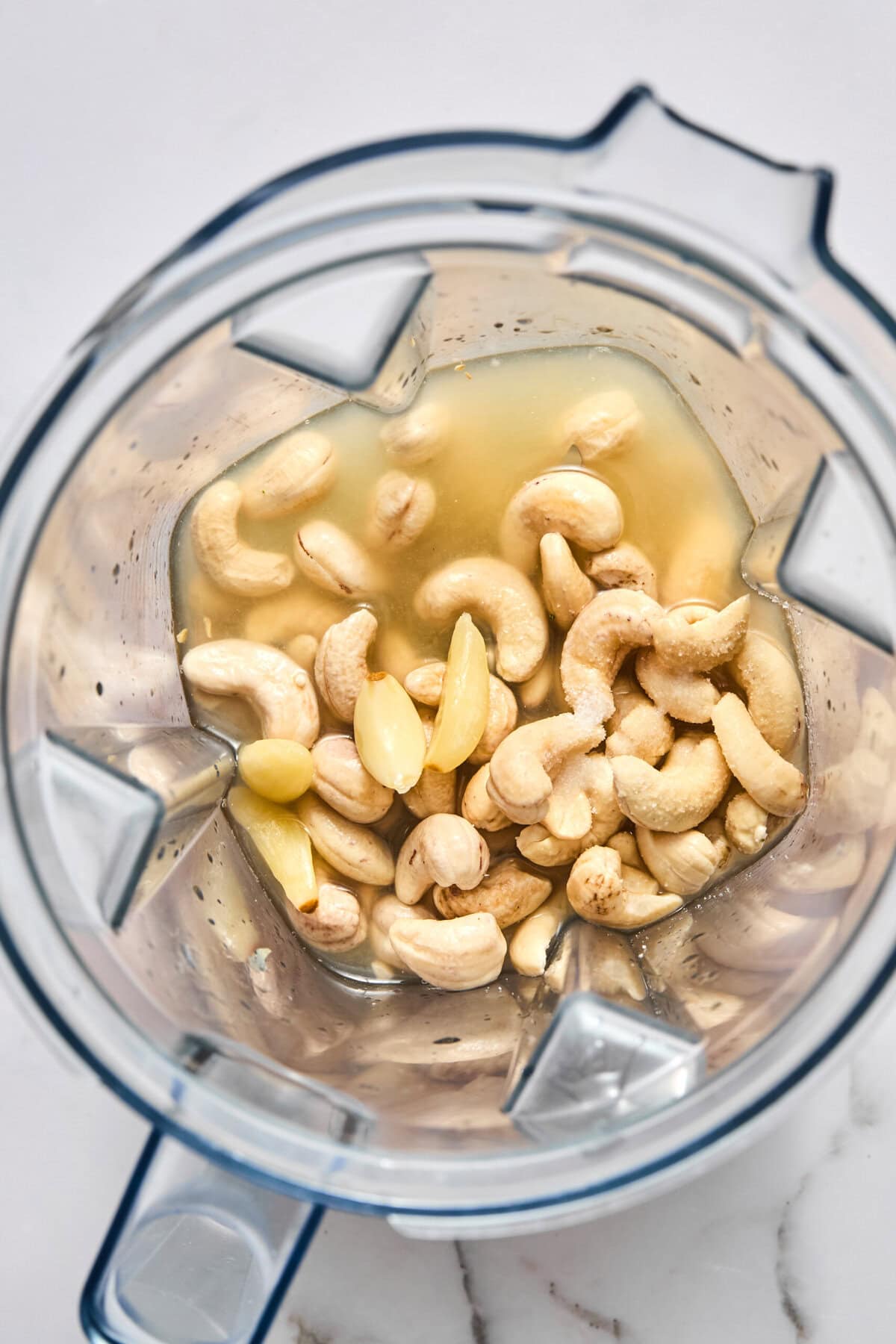 Raw cashews, garlic cloves, and water sit in a blender, ready to be mixed. The blender is viewed from above on a white surface.