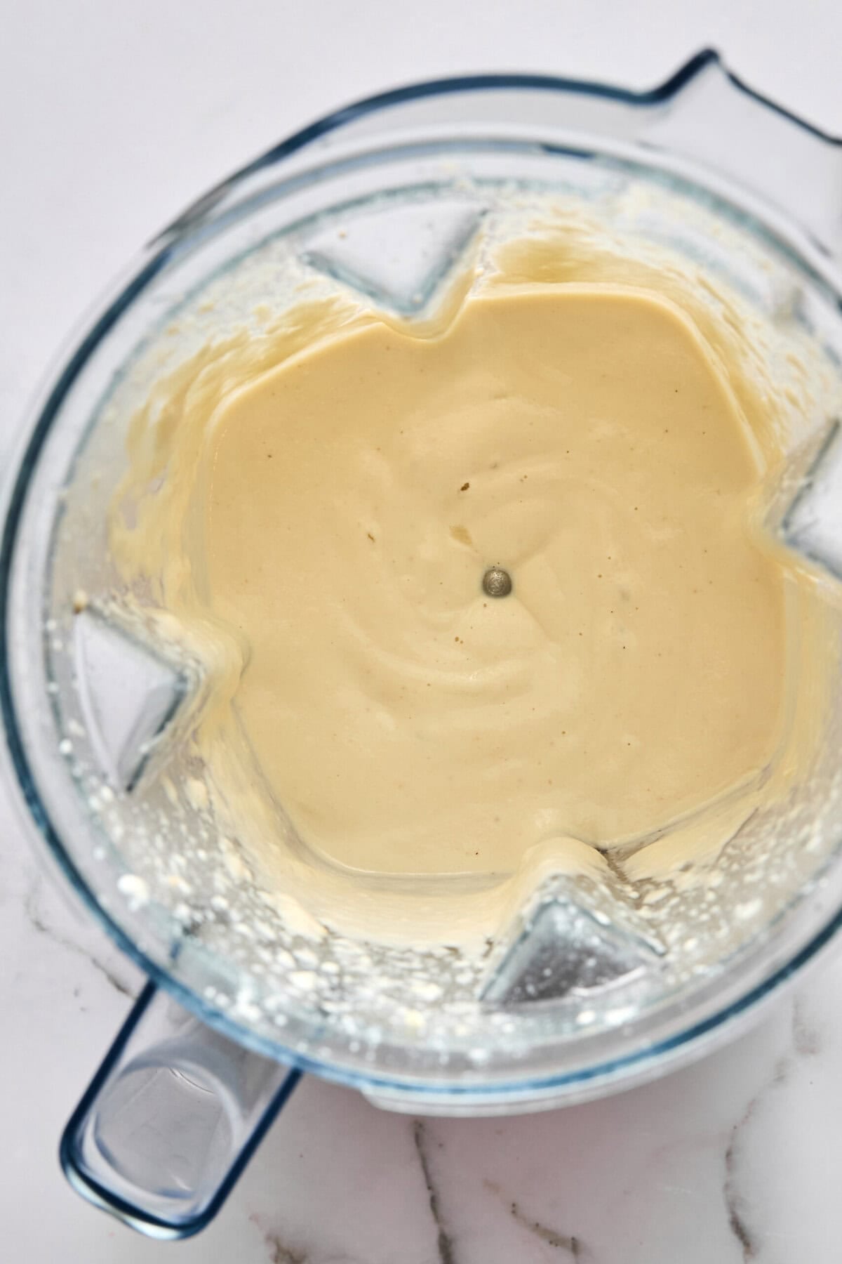 A top-down view of a blender filled with creamy, light beige batter or sauce, placed on a white marble surface.