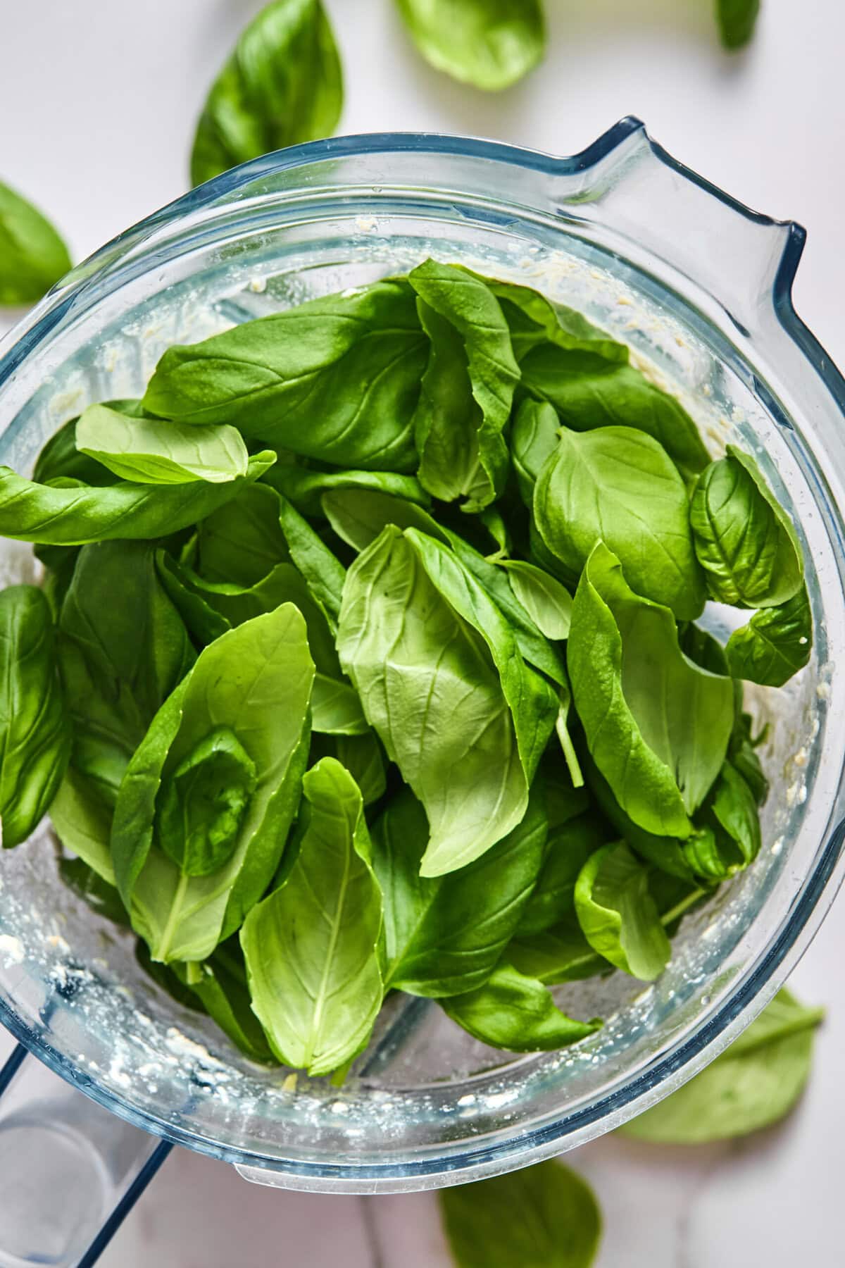 Fresh basil leaves in a food processor bowl, ready to be blended. Some small pieces of basil and other ingredients are visible at the bottom. A few more basil leaves are scattered on the white surface nearby.