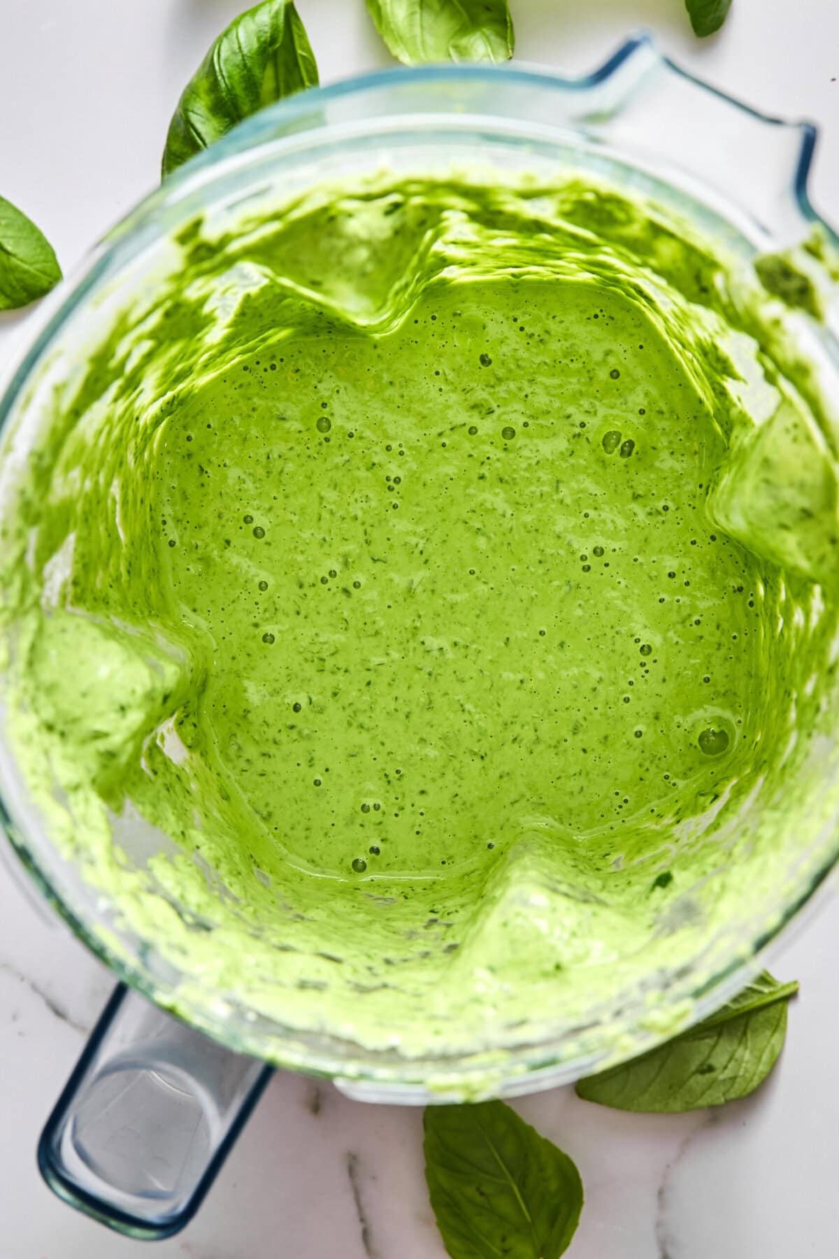 A top-down view of a blender filled with bright green sauce, likely pesto, surrounded by fresh basil leaves on a white surface.