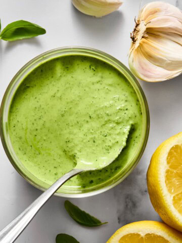 A bowl of creamy green sauce with a spoon, surrounded by fresh basil leaves, garlic cloves, and lemon halves on a white surface.