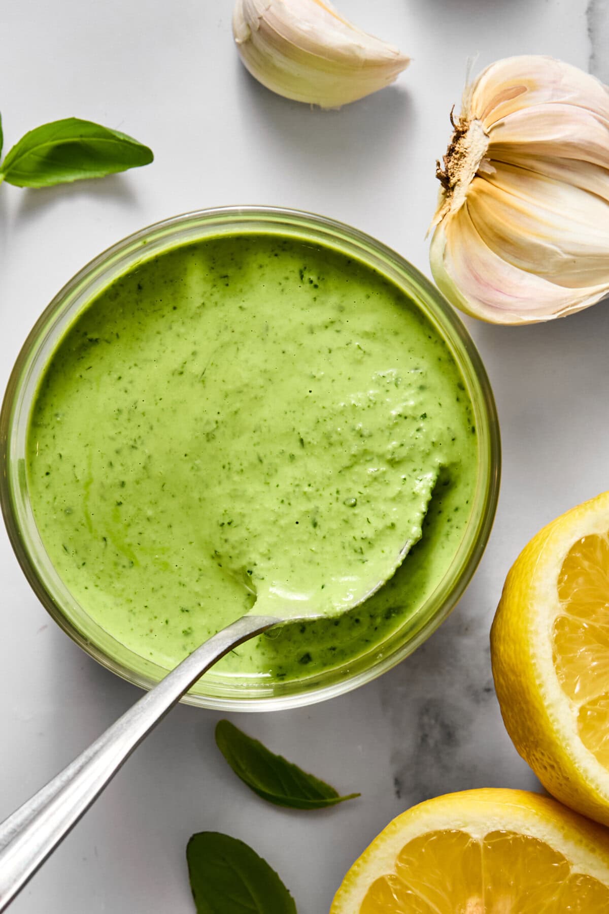 A bowl of creamy green sauce with a spoon, surrounded by fresh basil leaves, garlic cloves, and lemon halves on a white surface.