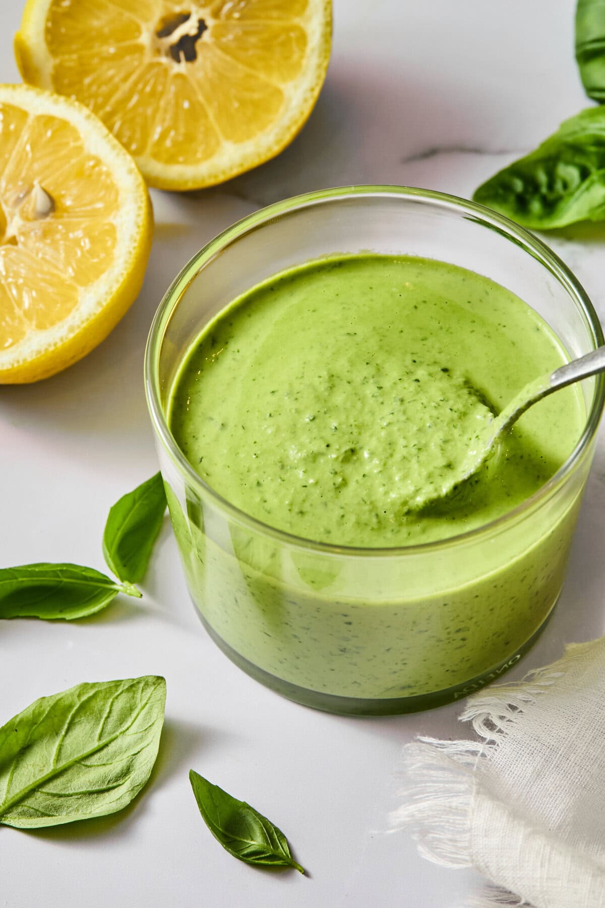 A glass bowl of creamy green sauce with a spoon, surrounded by fresh basil leaves and a halved lemon on a light surface.