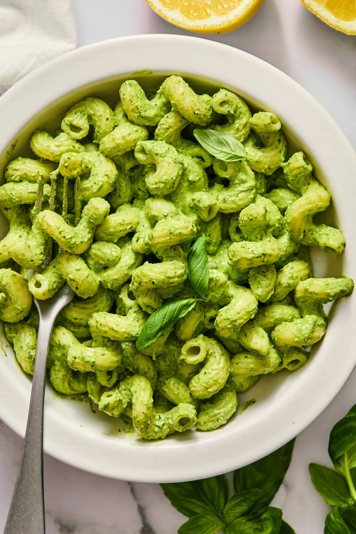 A white bowl filled with pasta coated in green pesto sauce, garnished with fresh basil leaves, and a fork placed inside. Lemon slices and basil leaves are nearby on a marble surface.