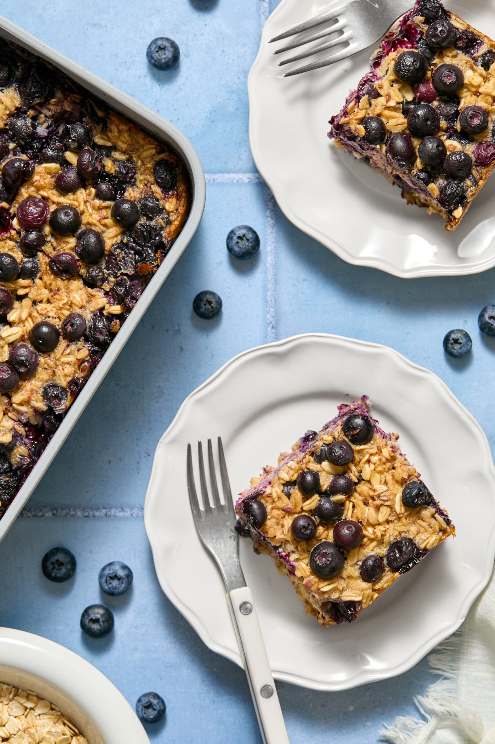 Overhead view of blueberry baked oats bars topped with fresh blueberries on white plates, with forks and loose blueberries scattered on a blue surface.