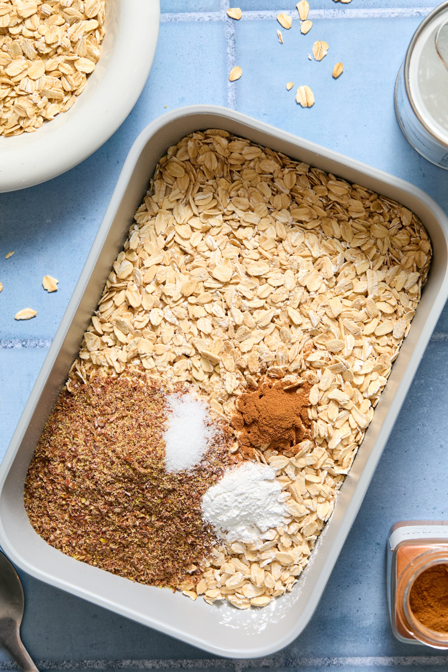 A rectangular pan filled with blueberry baked oats, ground flaxseed, salt, baking powder, and cinnamon sits on a blue tiled surface. A bowl of oats, glass jar, and spice jar are nearby.