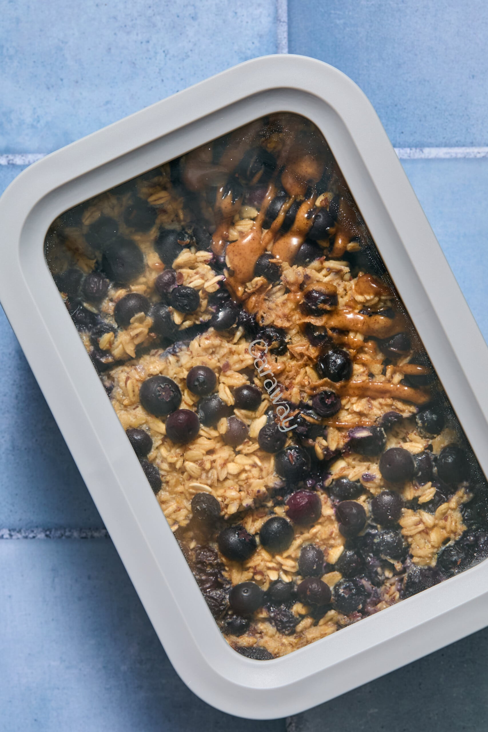 A rectangular container with a clear lid shows a delicious blueberry baked oats dish topped with fresh blueberries, sitting on a blue tiled surface.
