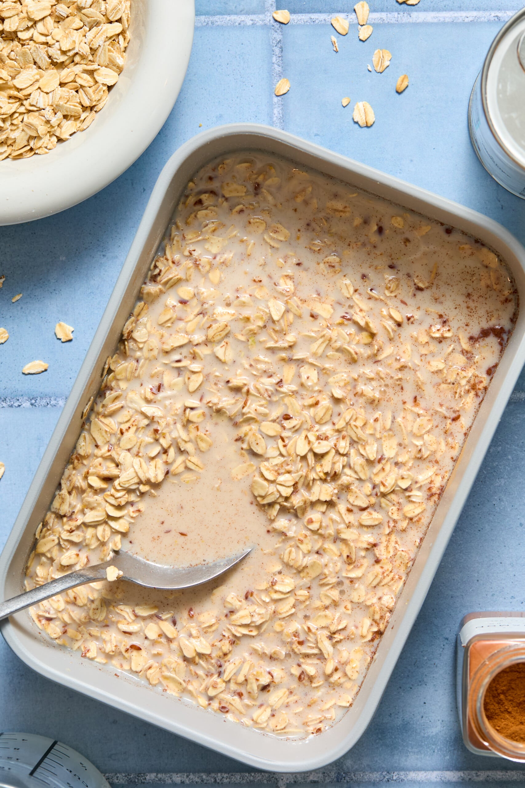 A baking dish filled with blueberry baked oats and milk mixture sits on a blue surface, with a spoon resting inside. Scattered oats, a bowl of oats, and a jar are visible nearby.