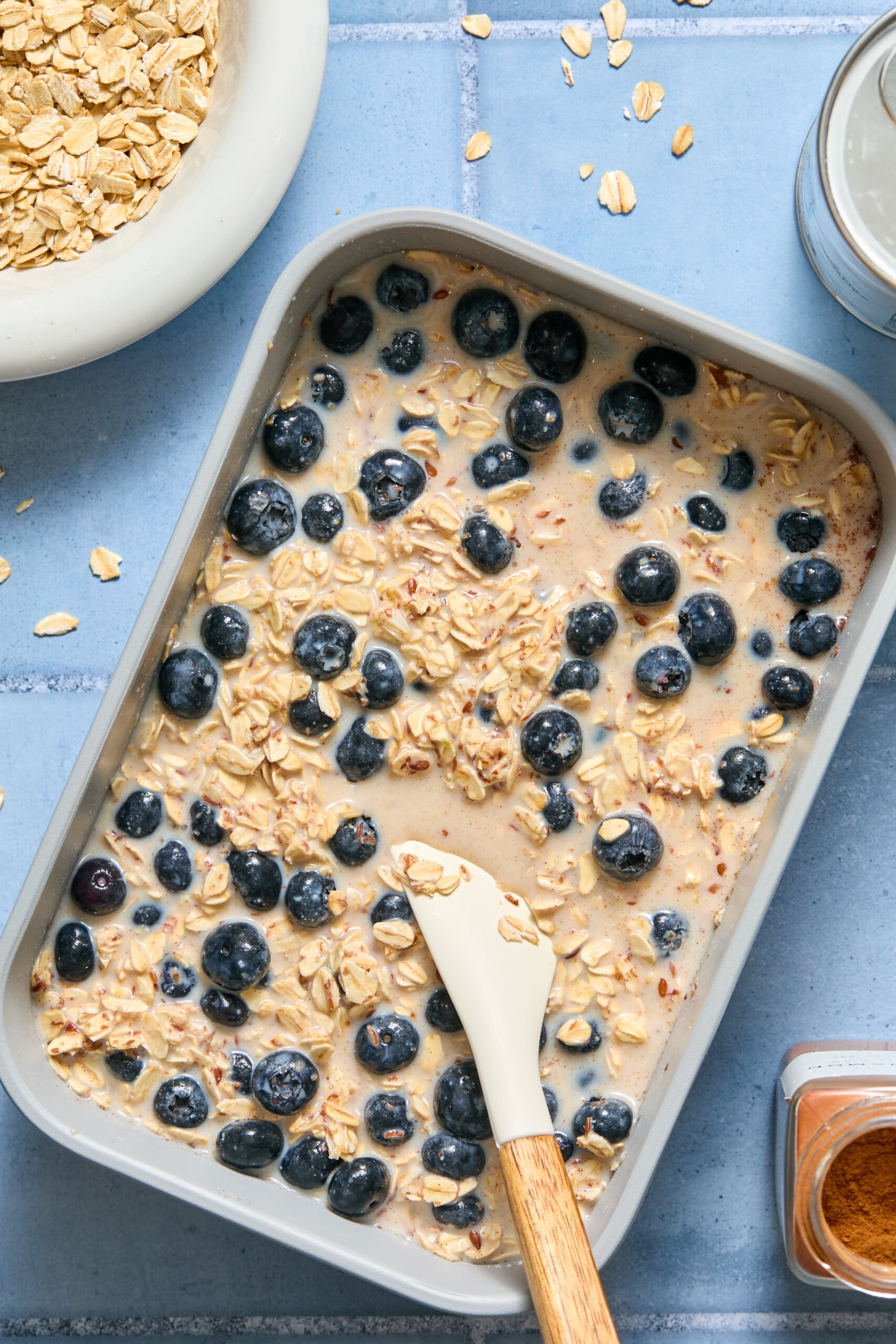 A baking dish filled with blueberry baked oats, fresh blueberries, and a creamy liquid mixture sits on a blue surface, with a spatula resting inside. Nearby are a bowl of oats, spilled oats, and a jar of cinnamon.