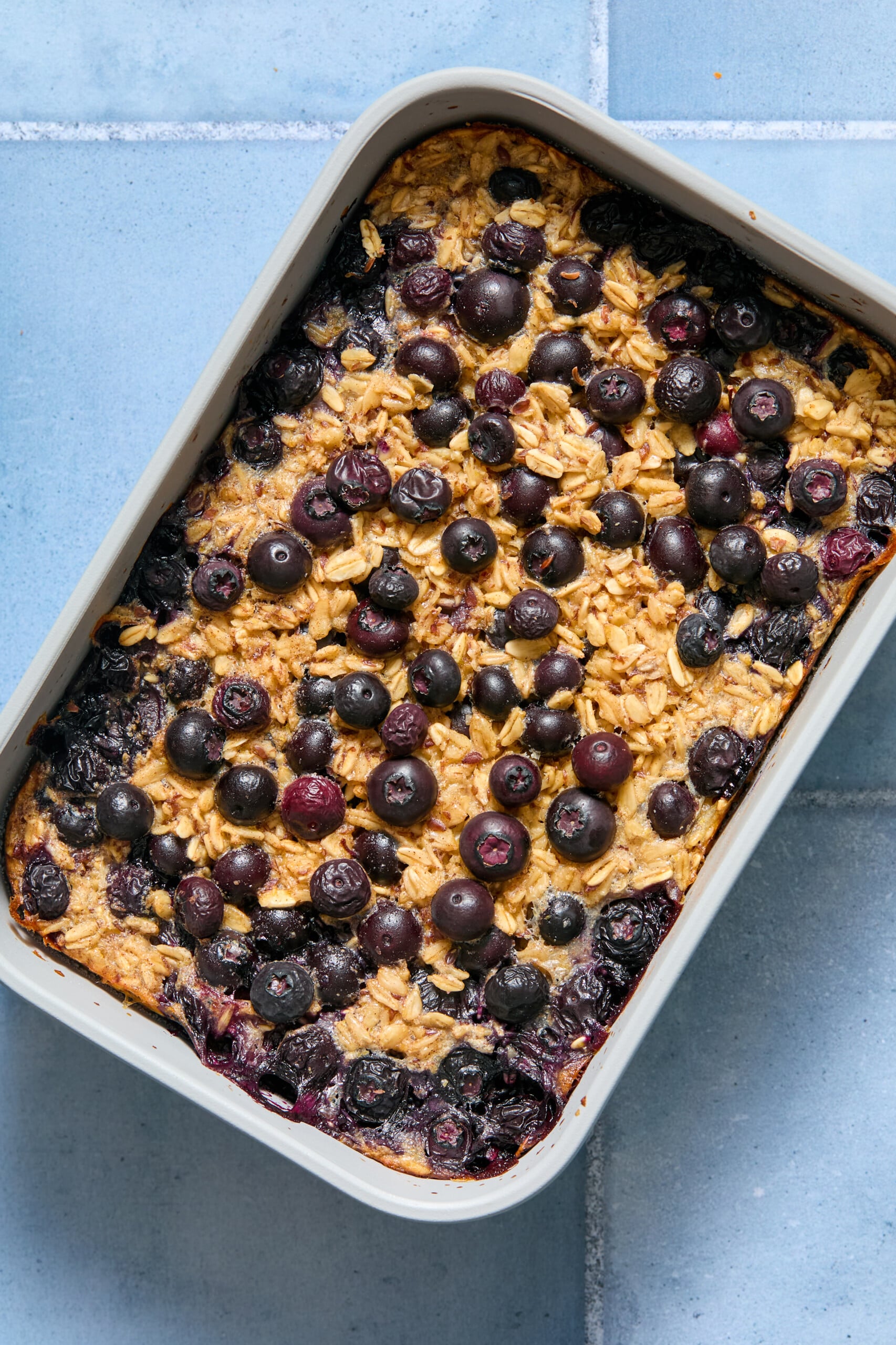 A blueberry baked oats dish, topped with fresh blueberries, sits in a rectangular baking pan on a blue tiled surface.