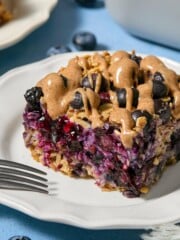 A slice of blueberry baked oats with plump blueberries and a drizzle of nut butter sits on a white plate, with a fork nearby and loose blueberries scattered on the blue table.