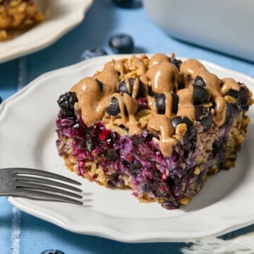 A slice of blueberry baked oats with plump blueberries and a drizzle of nut butter sits on a white plate, with a fork nearby and loose blueberries scattered on the blue table.