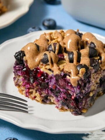 A slice of blueberry baked oats with plump blueberries and a drizzle of nut butter sits on a white plate, with a fork nearby and loose blueberries scattered on the blue table.