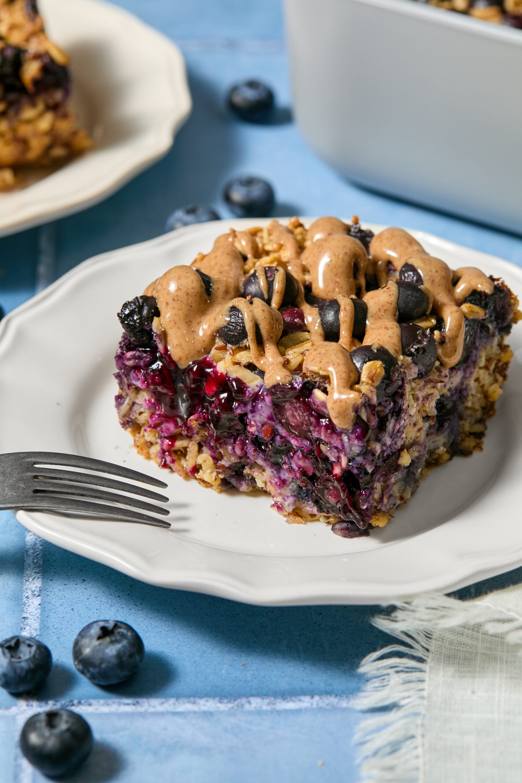 A slice of blueberry baked oats with plump blueberries and a drizzle of nut butter sits on a white plate, with a fork nearby and loose blueberries scattered on the blue table.