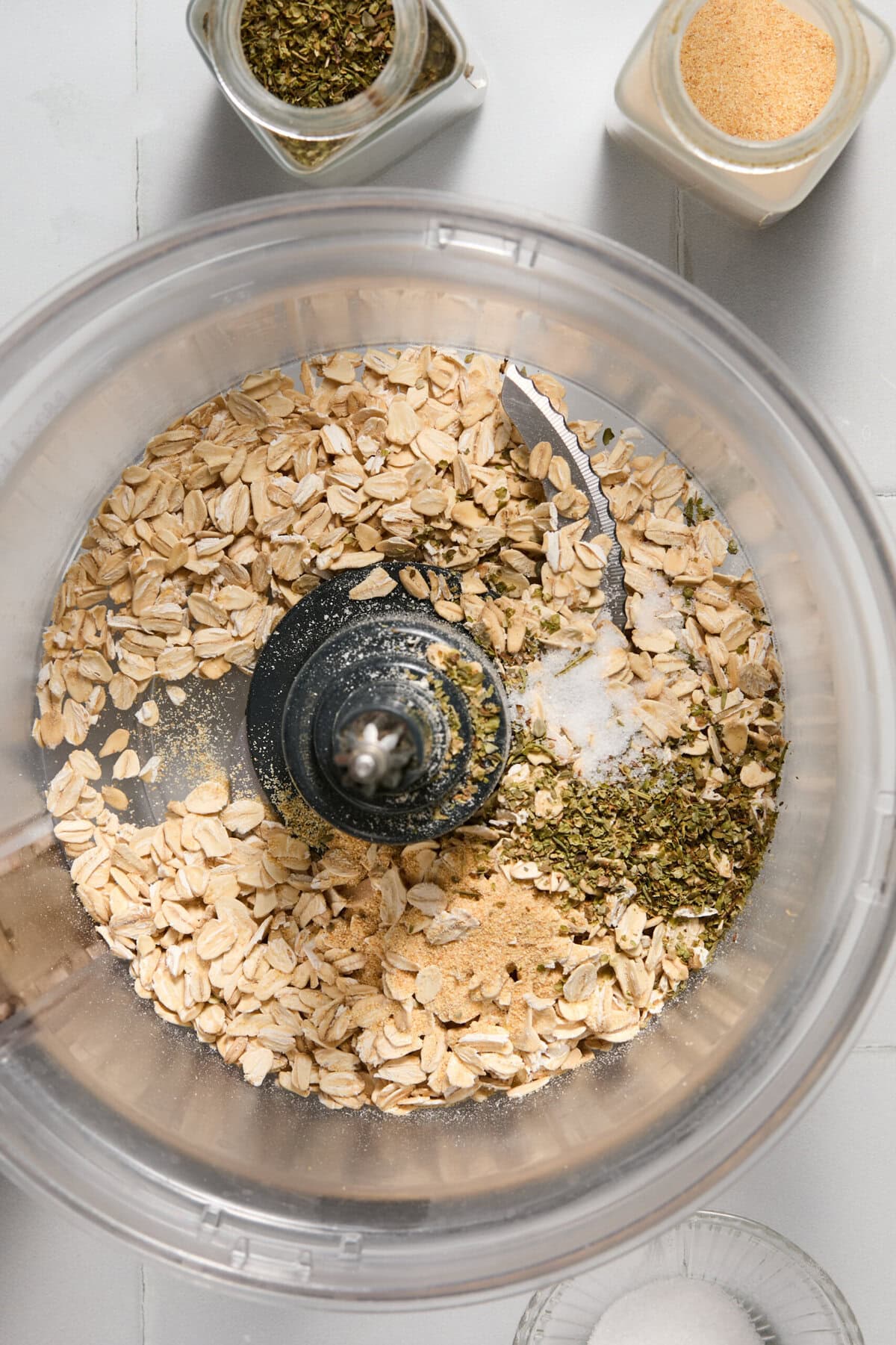 A food processor bowl contains rolled oats, dried herbs, salt, and garlic powder, with spice jars and a small bowl of salt nearby on a light countertop.