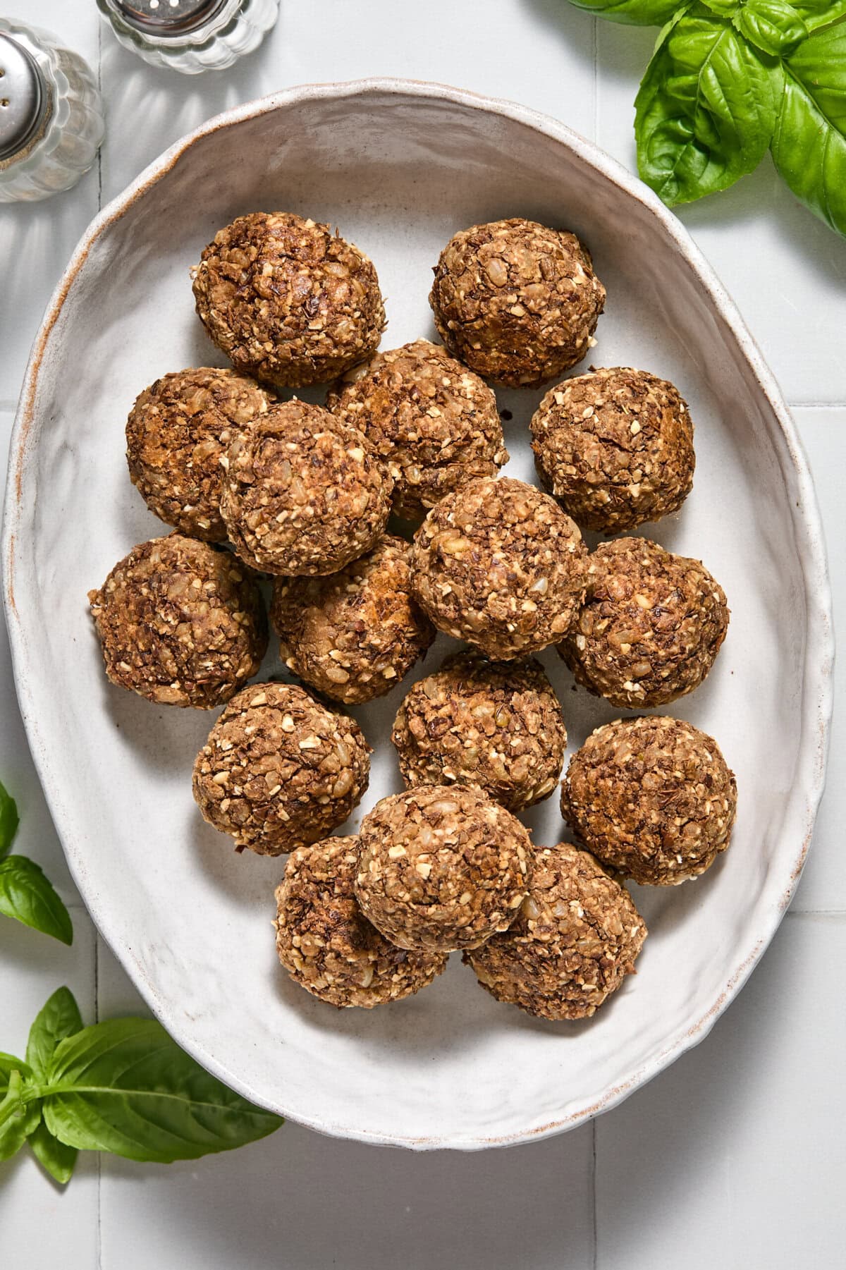 A white oval platter holds a pile of round, brown, oat-filled baked balls. Fresh basil leaves and salt and pepper shakers are nearby on a light surface.