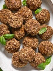 A white oval plate with round, brown oat and nut balls, garnished with fresh green basil leaves, on a light-colored table with some basil leaves around.