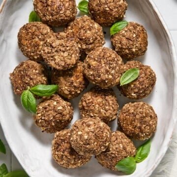A white oval plate with round, brown oat and nut balls, garnished with fresh green basil leaves, on a light-colored table with some basil leaves around.