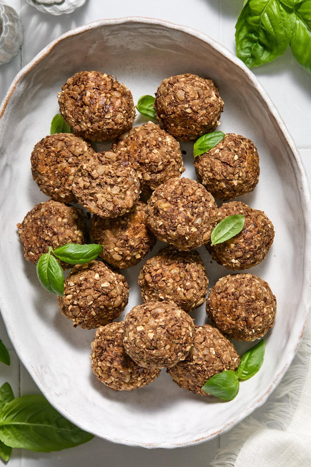 A white oval plate with round, brown oat and nut balls, garnished with fresh green basil leaves, on a light-colored table with some basil leaves around.