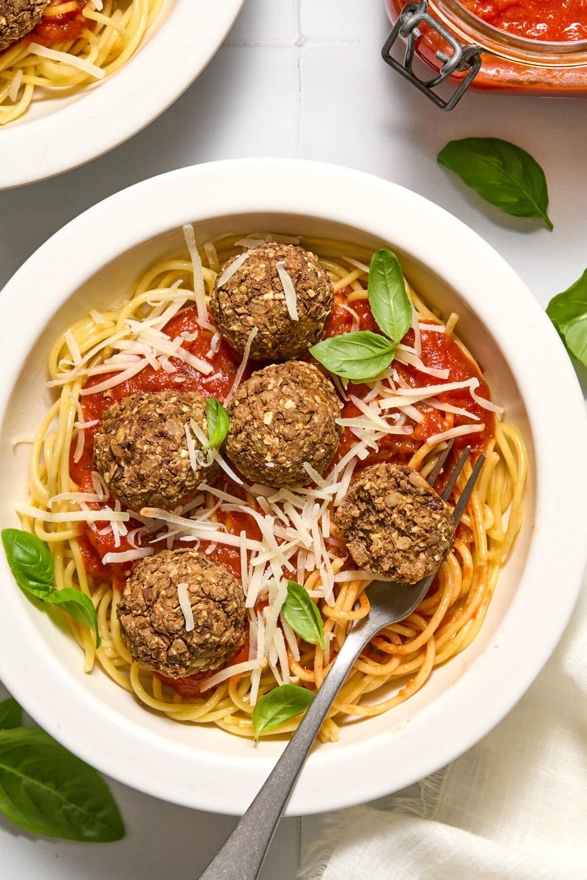 A bowl of spaghetti topped with tomato sauce, plant-based meatballs, shredded cheese, and fresh basil leaves. A fork rests in the bowl.