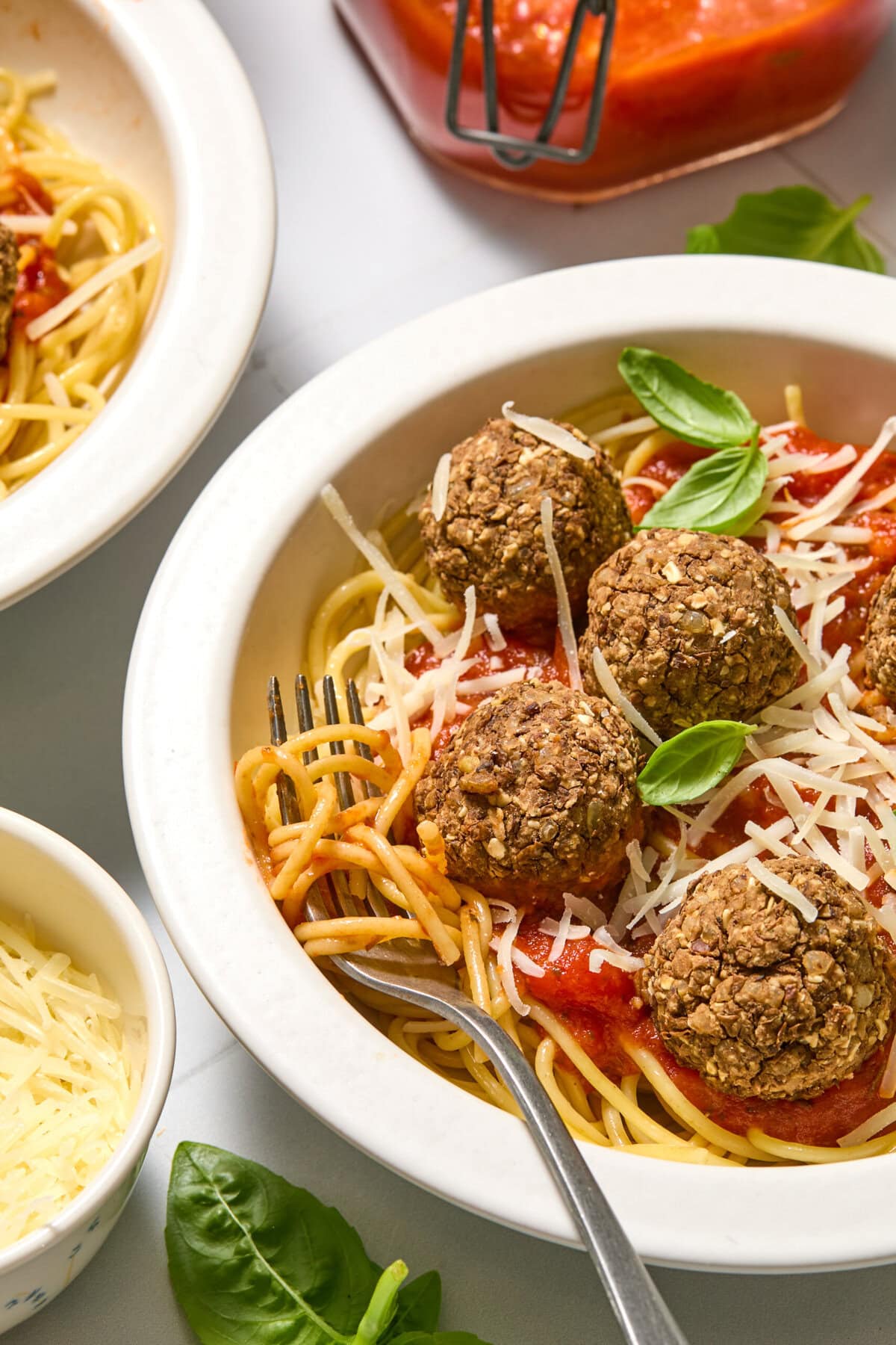 A bowl of spaghetti topped with tomato sauce, hearty Plant-Based Meatballs, shredded cheese, and fresh basil. A fork rests in the bowl; another bowl and a jar of sauce are nearby.