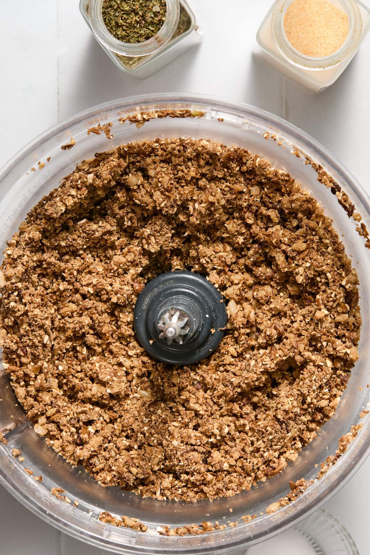 A food processor bowl filled with a coarse, brown, crumbly mixture, with jars of dried herbs and garlic powder nearby on a white surface.
