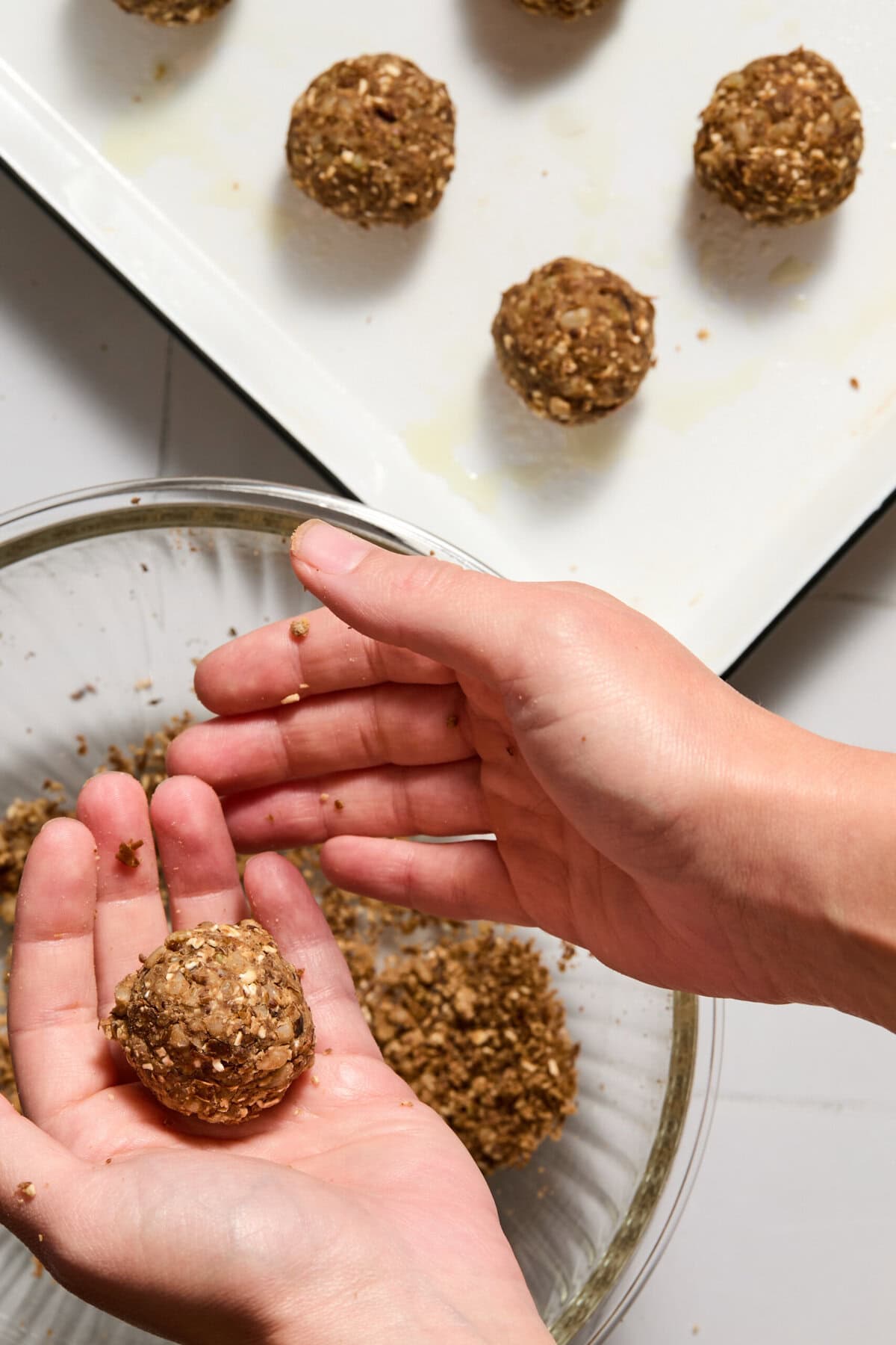 Two hands shape an oat and seed mixture into a ball over a bowl. Several finished balls are arranged on a white tray nearby.
