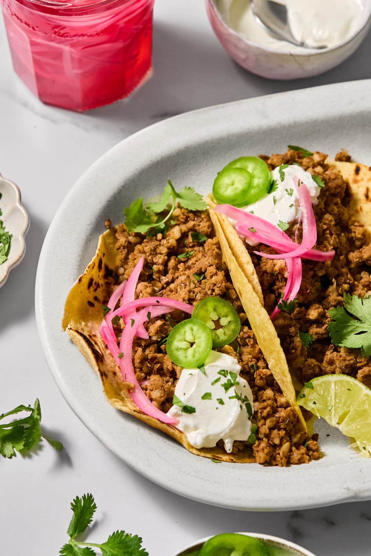 A plate with two ground beef tacos topped with sour cream, pickled onions, jalapeño slices, cilantro, and a lime wedge. A glass of pink drink and small bowls of sauce are in the background.
