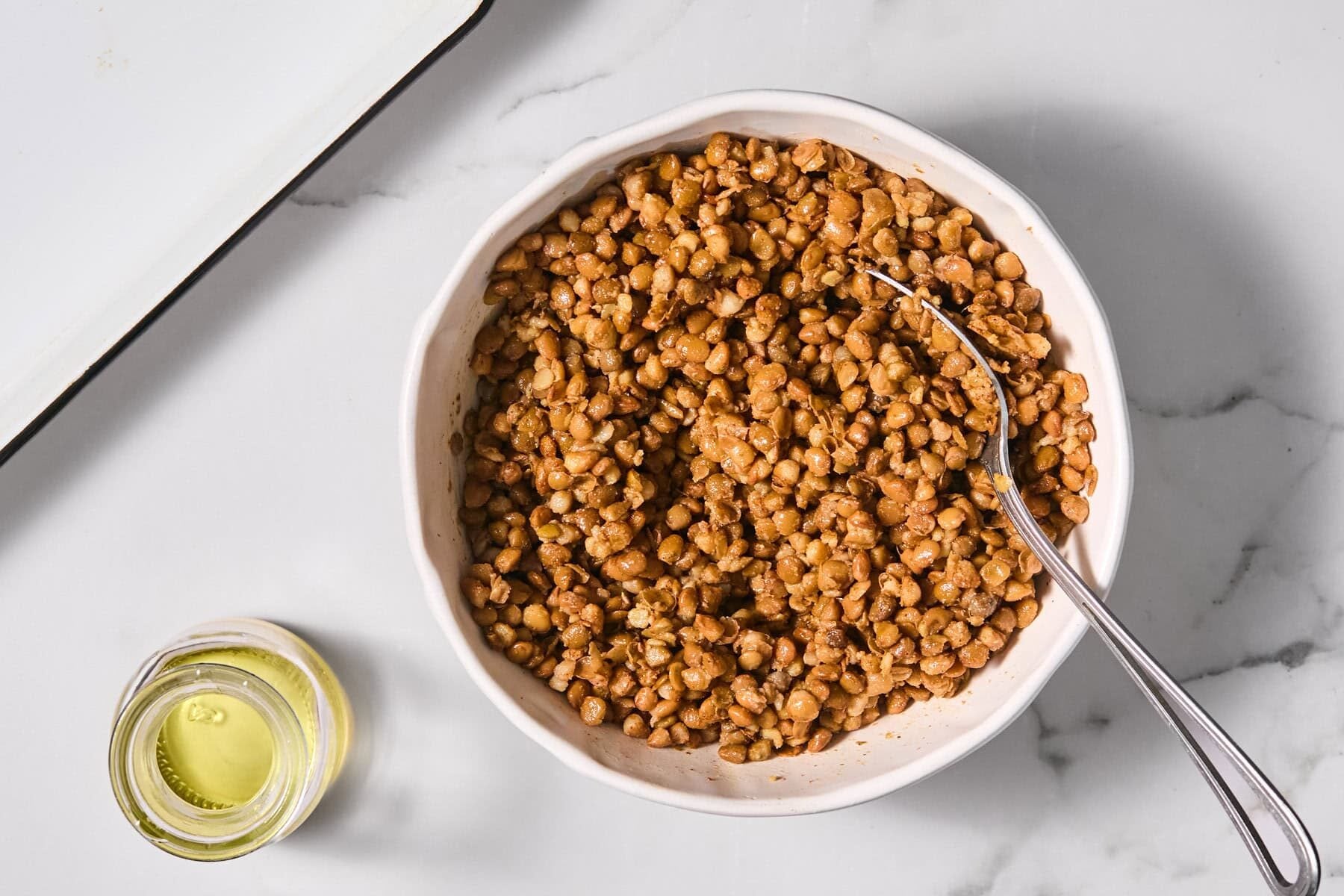A white bowl filled with mashed chickpeas and spices, with a fork inside. A small glass jar with olive oil is placed nearby on a white marble surface.