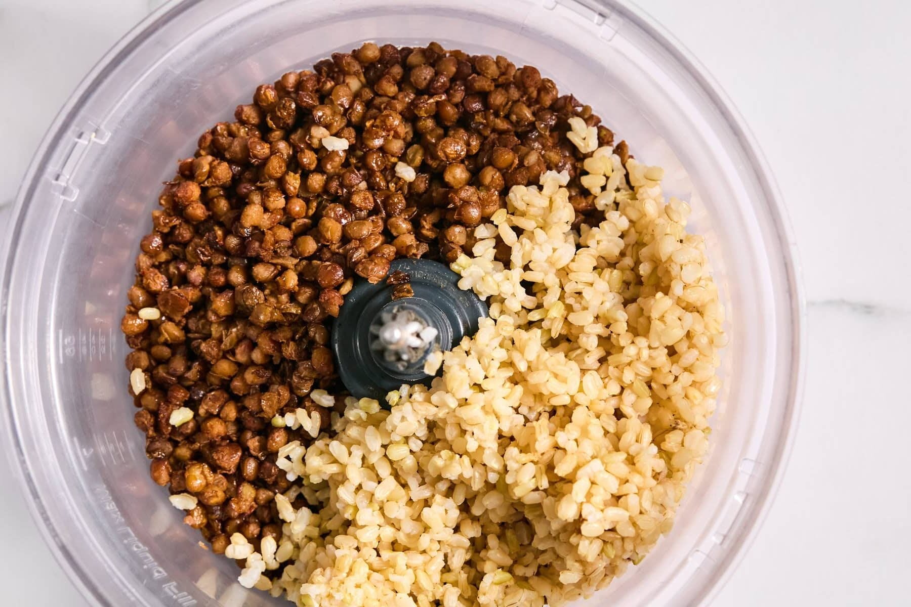 A food processor bowl filled with cooked brown lentils on one side and cooked brown rice on the other, ready to be blended.