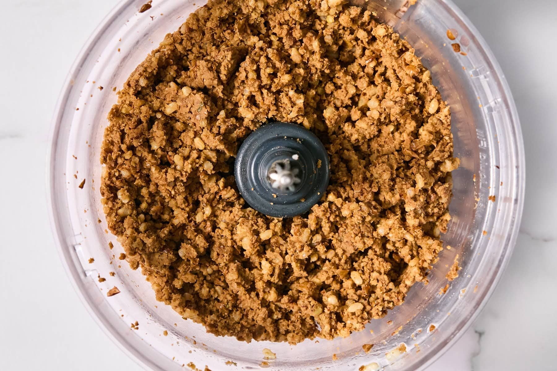 Crumbled brown mixture with visible nuts in a food processor bowl, viewed from above on a white surface.