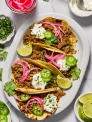 Three tacos filled with seasoned ground meat, topped with sour cream, pickled onions, jalapeño slices, cilantro, and lime wedges on a white plate. Bowls of toppings and lime slices are nearby.
