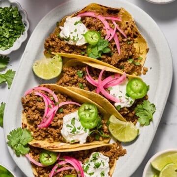 Three tacos filled with seasoned ground meat, topped with sour cream, pickled onions, jalapeño slices, cilantro, and lime wedges on a white plate. Bowls of toppings and lime slices are nearby.