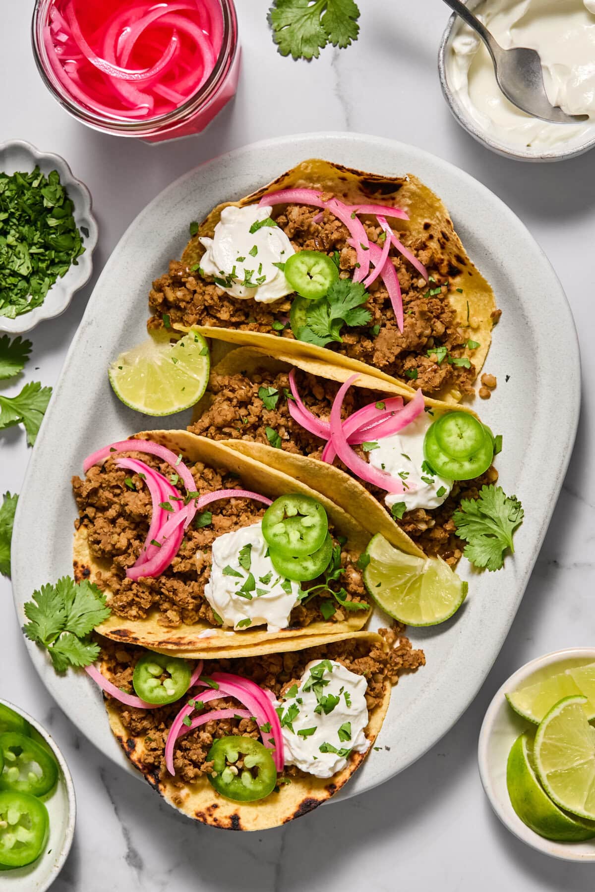 Three tacos filled with seasoned ground meat, topped with sour cream, pickled onions, jalape&ntilde;o slices, cilantro, and lime wedges on a white plate. Bowls of toppings and lime slices are nearby.