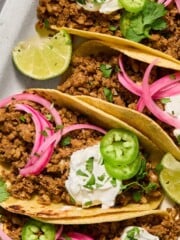 Three tacos filled with seasoned ground meat, topped with sour cream, sliced jalapeños, pickled red onions, chopped cilantro, and lime wedges on the side, served on a white plate.