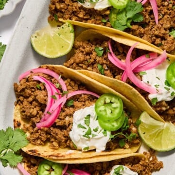 Three tacos filled with seasoned ground meat, topped with sour cream, sliced jalapeños, pickled red onions, chopped cilantro, and lime wedges on the side, served on a white plate.
