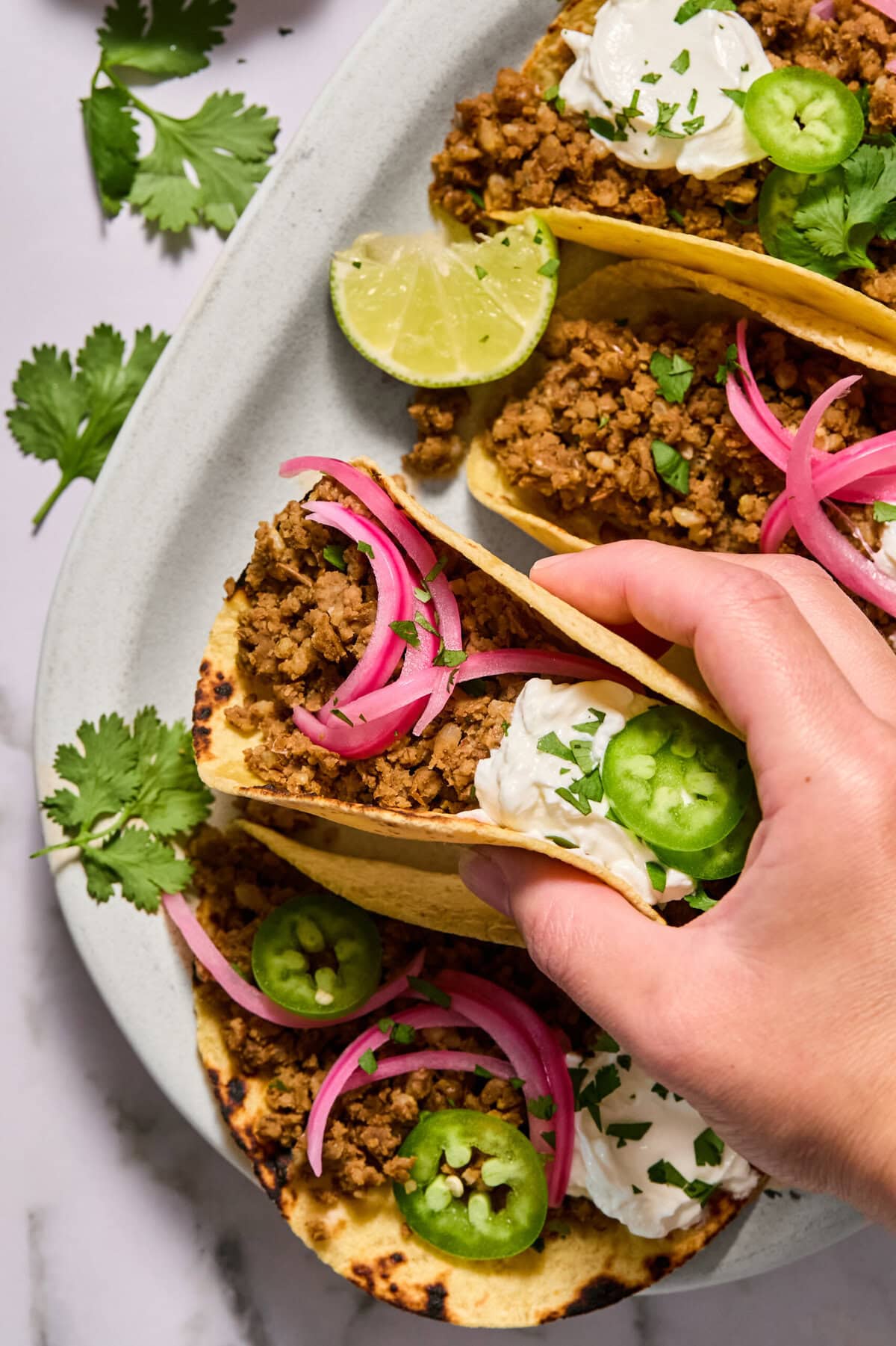 A hand picks up a taco filled with ground meat, sour cream, pickled onions, jalapeño slices, and cilantro. Two more tacos with similar toppings sit on a plate beside lime wedges and fresh cilantro.