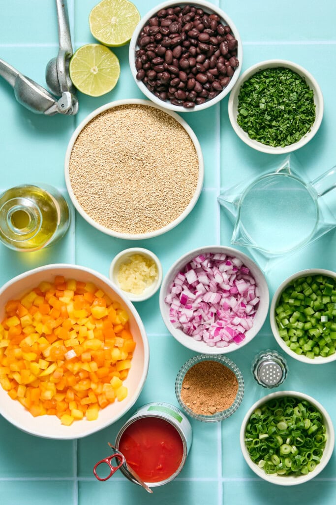 Bowls of black beans, quinoa, chopped yellow bell peppers, red onion, green onion, cilantro, jalapeño, garlic, and spices, with a halved lime, olive oil, canned tomatoes, and a pitcher of water on a blue surface.