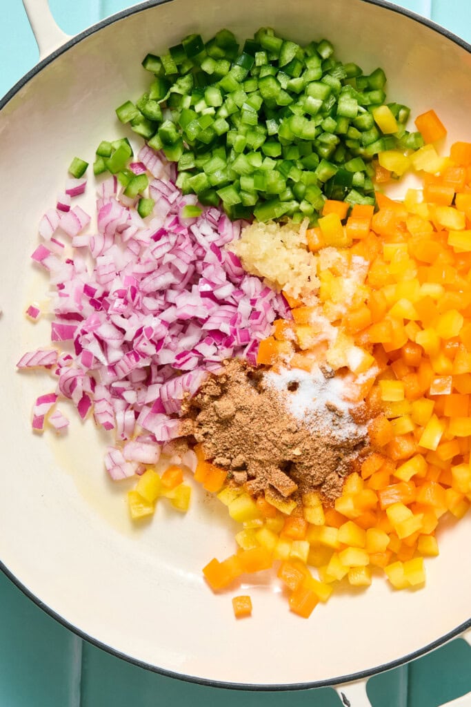 Chopped green bell pepper, yellow bell pepper, and red onion, with minced garlic, ground spices, and salt in a white pan on a blue surface, ready to be cooked.