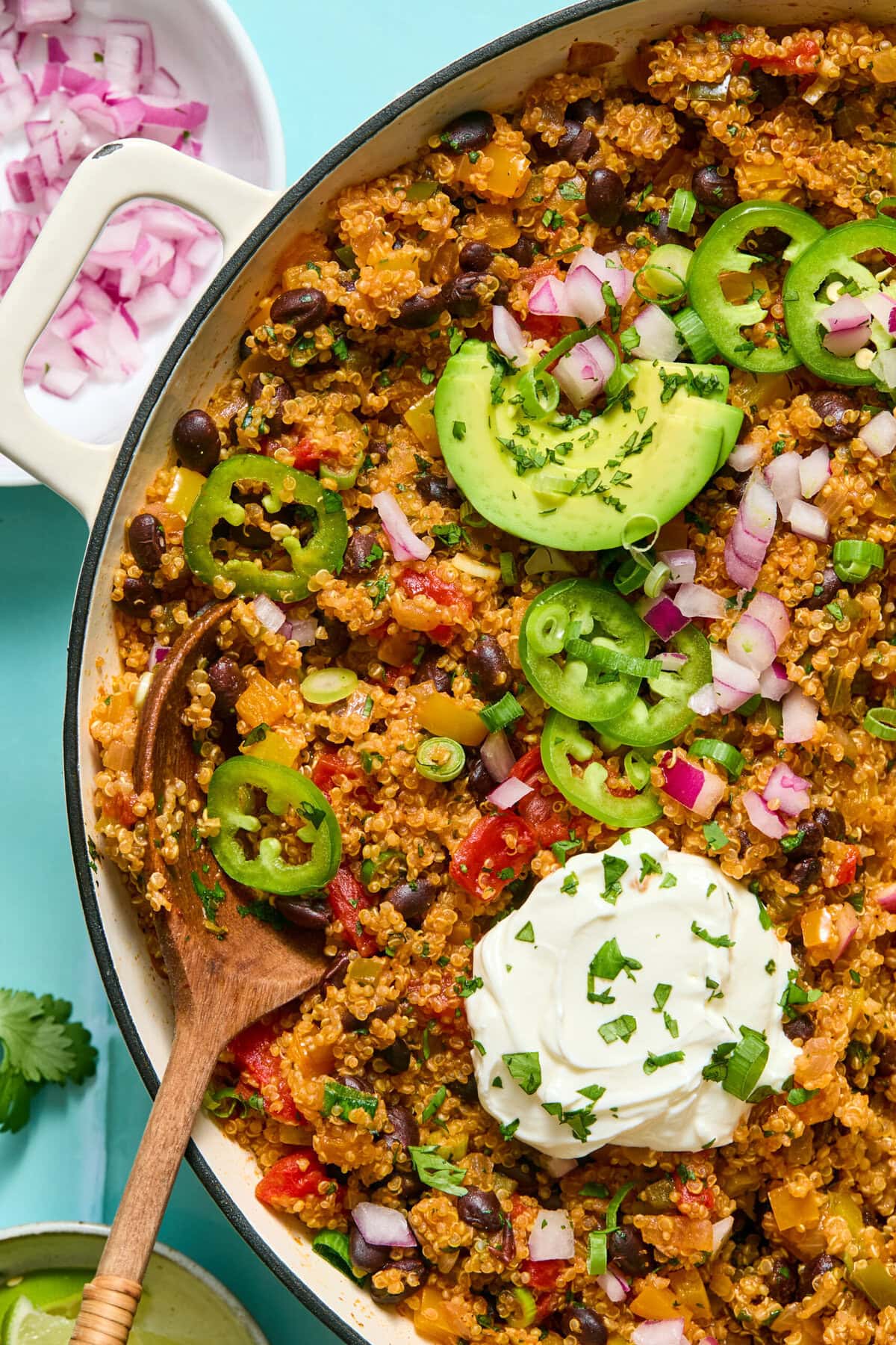 A skillet filled with quinoa, black beans, tomatoes, sliced jalapeños, avocado, chopped red onion, and a dollop of sour cream, garnished with cilantro. A wooden spoon rests inside the skillet; a bowl of onions is nearby.