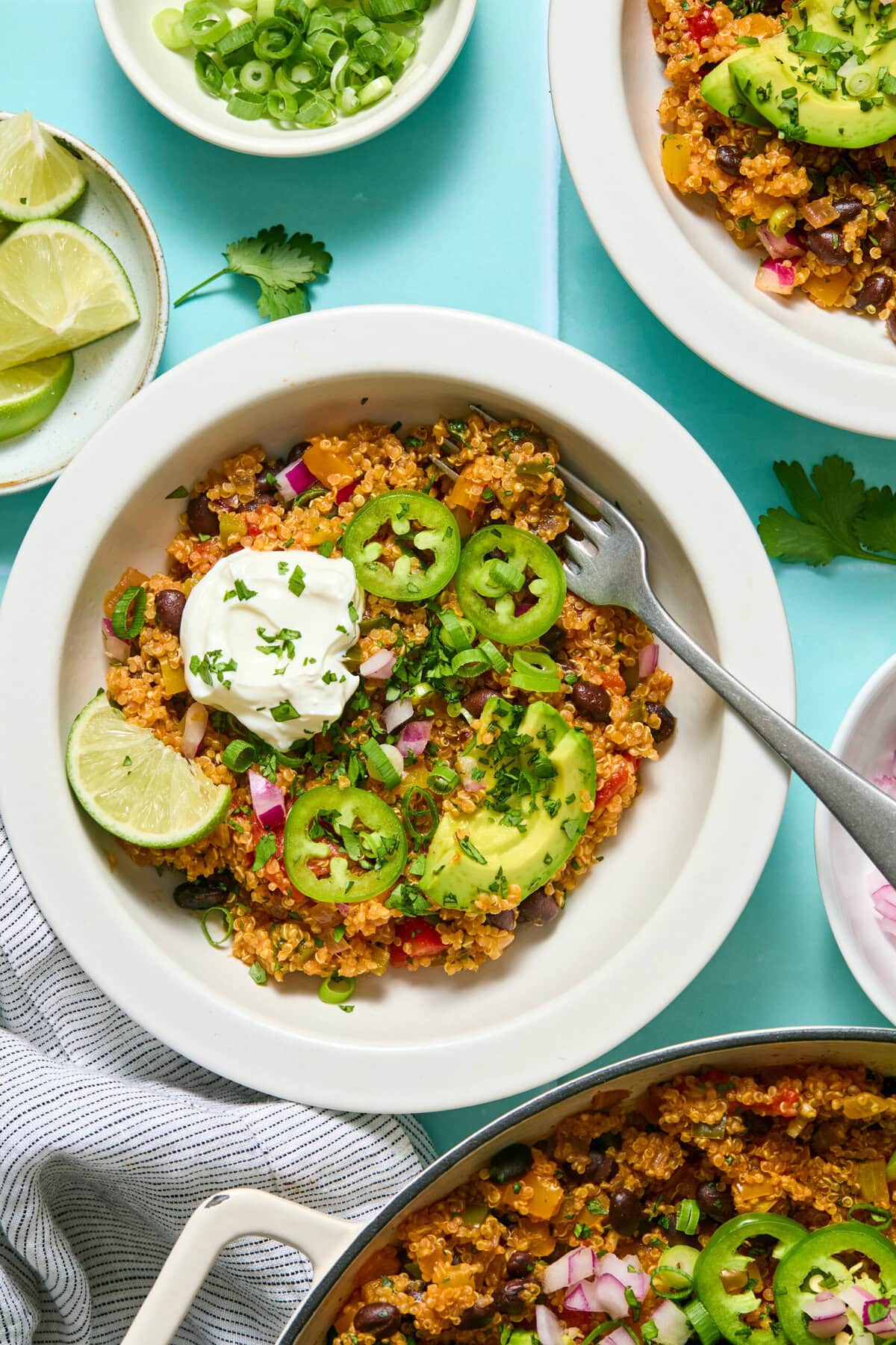 A bowl of quinoa and black bean salad topped with avocado slices, jalapeños, sour cream, lime wedge, and chopped herbs, with a fork, green onions, and lime on the side.